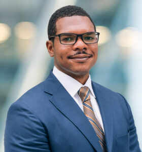A man wearing glasses, a blue suit, a white shirt, and a striped tie smiles slightly while standing against a blurred background with soft lights.