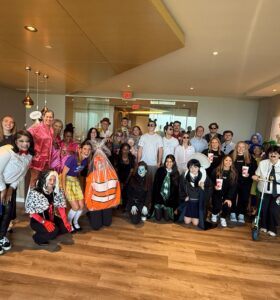 A large group of people in colorful and creative costumes pose together in an office lobby with wooden floors and modern decor, smiling and celebrating a festive occasion.