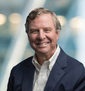 A middle-aged man in a navy suit and white shirt smiles at the camera. The background is blurred with soft light and abstract shapes, suggesting an indoor office setting.