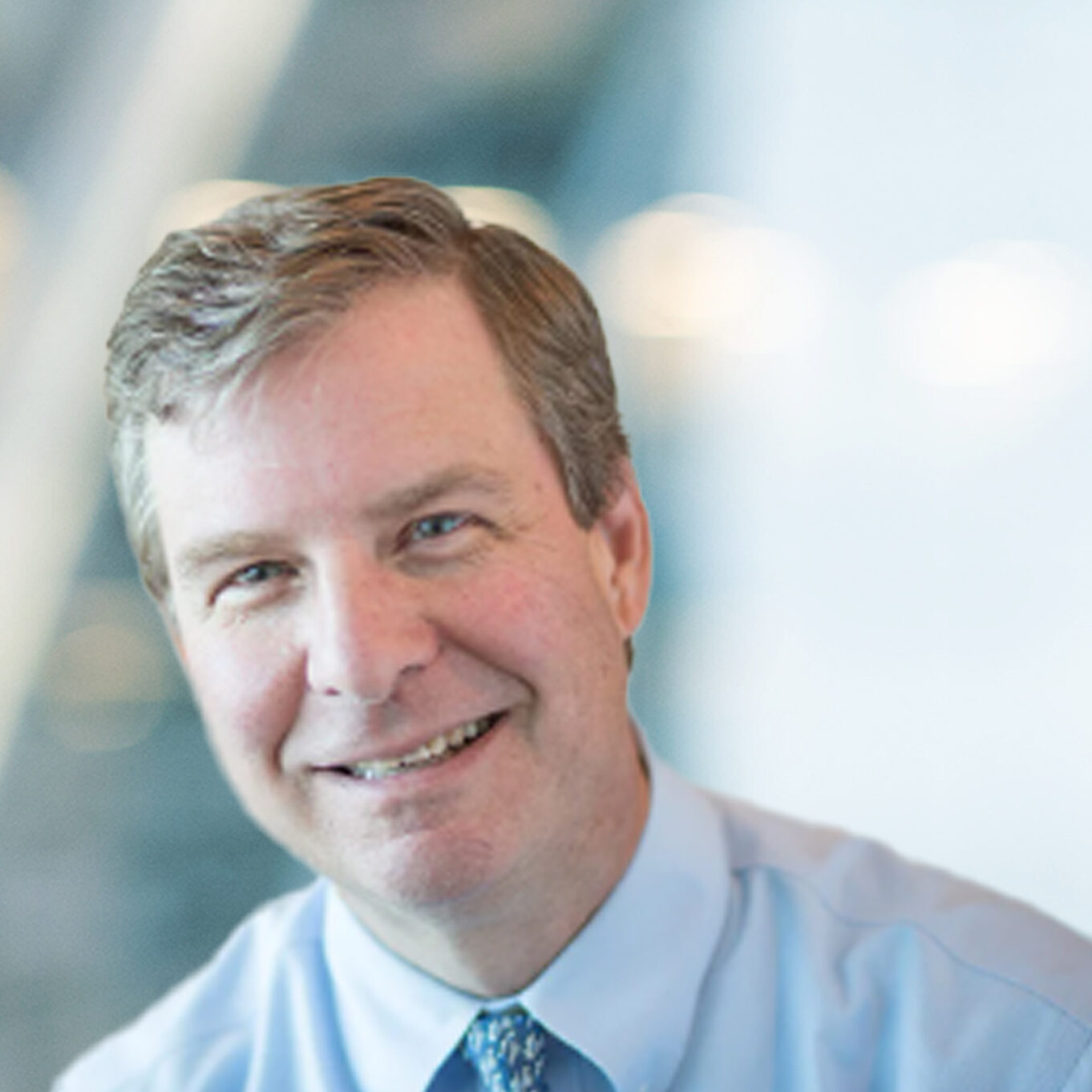 A middle-aged man in a dress shirt and patterned tie smiles at the camera, with a blurred modern office background and soft lighting.
