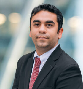 A man in a black suit, white shirt, and red tie poses for a professional headshot against a blurred background of blue and white tones. He has short dark hair and a neutral expression.