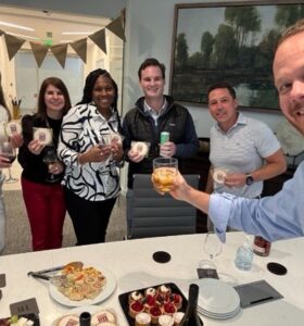 Six people stand around a table with drinks and snacks, smiling at the camera. One person holds up a drink in a toast. The scene appears festive, with food, desserts, and a decorated banner in the background.