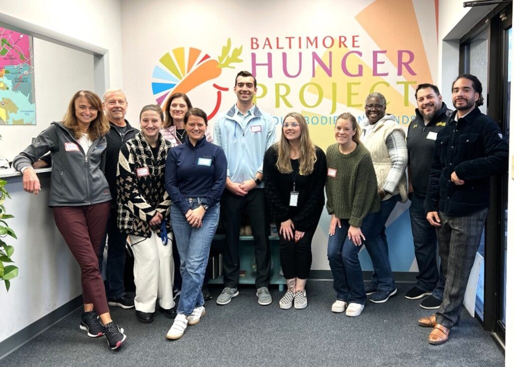 A group of twelve smiling adults stands together in front of a colorful Baltimore Hunger Project sign in an indoor office, posing for a group photo. Some are wearing name tags and casual clothes.