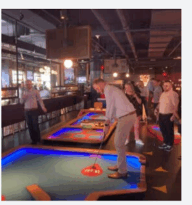 A group of adults play mini golf indoors on brightly lit, colorful courses. One man is about to putt while others watch and socialize in a lively, modern venue with exposed ceilings and hanging lights.