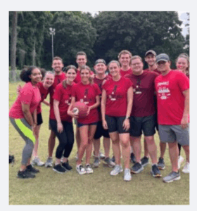 A group of twelve smiling people, mostly wearing matching red shirts, pose together outdoors on grass. One person holds a ball, suggesting a team sports event. Trees and light poles are visible in the background.