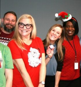 Six adults stand together indoors, smiling at the camera. One person wears a Christmas-themed T-shirt, another has a Santa hat headband, and one wears a festive sweater. The group appears cheerful and relaxed.