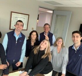 Six people, four men and two women, smile and pose together in an office setting around a table. Everyone appears friendly and cheerful, with a diploma framed on the wall behind them.