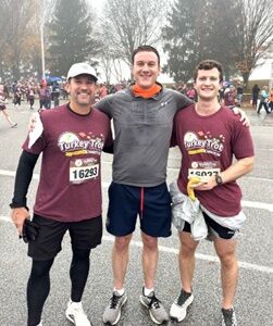 Three men stand smiling after a race, two wearing matching maroon “Turkey Trot” shirts and race bibs, and one in a gray jacket. Other runners and trees are visible in the foggy background.