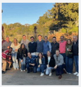 A group of sixteen adults, some standing and some kneeling, pose together outdoors on a sunny day with greenery and trees in the background. Everyone is smiling and dressed in casual clothing.