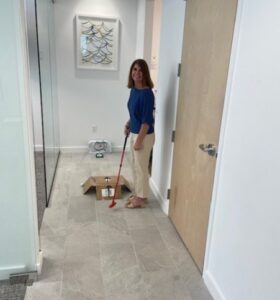 A woman stands in an office hallway, smiling while playing mini golf with a red putter. A cardboard box acts as a golf hole, and office decor including artwork and glass walls are visible in the background.