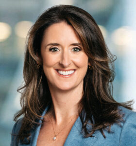 A woman with long brown hair wearing a light blue blazer and a necklace, smiling in front of a blurred, light-colored background.
