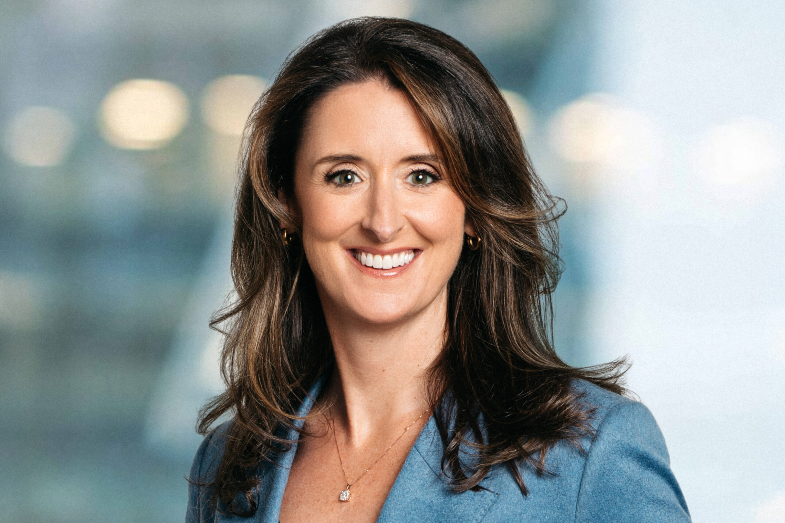 A woman with long brown hair wearing a light blue blazer and a necklace, smiling in front of a blurred, light-colored background.