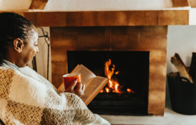 A person wrapped in a blanket sits by a lit fireplace, holding a book and a red mug, enjoying a cozy and relaxing moment indoors.