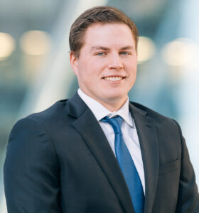 A young man in a dark suit, white shirt, and blue tie stands smiling in front of a blurred modern background with soft lighting.