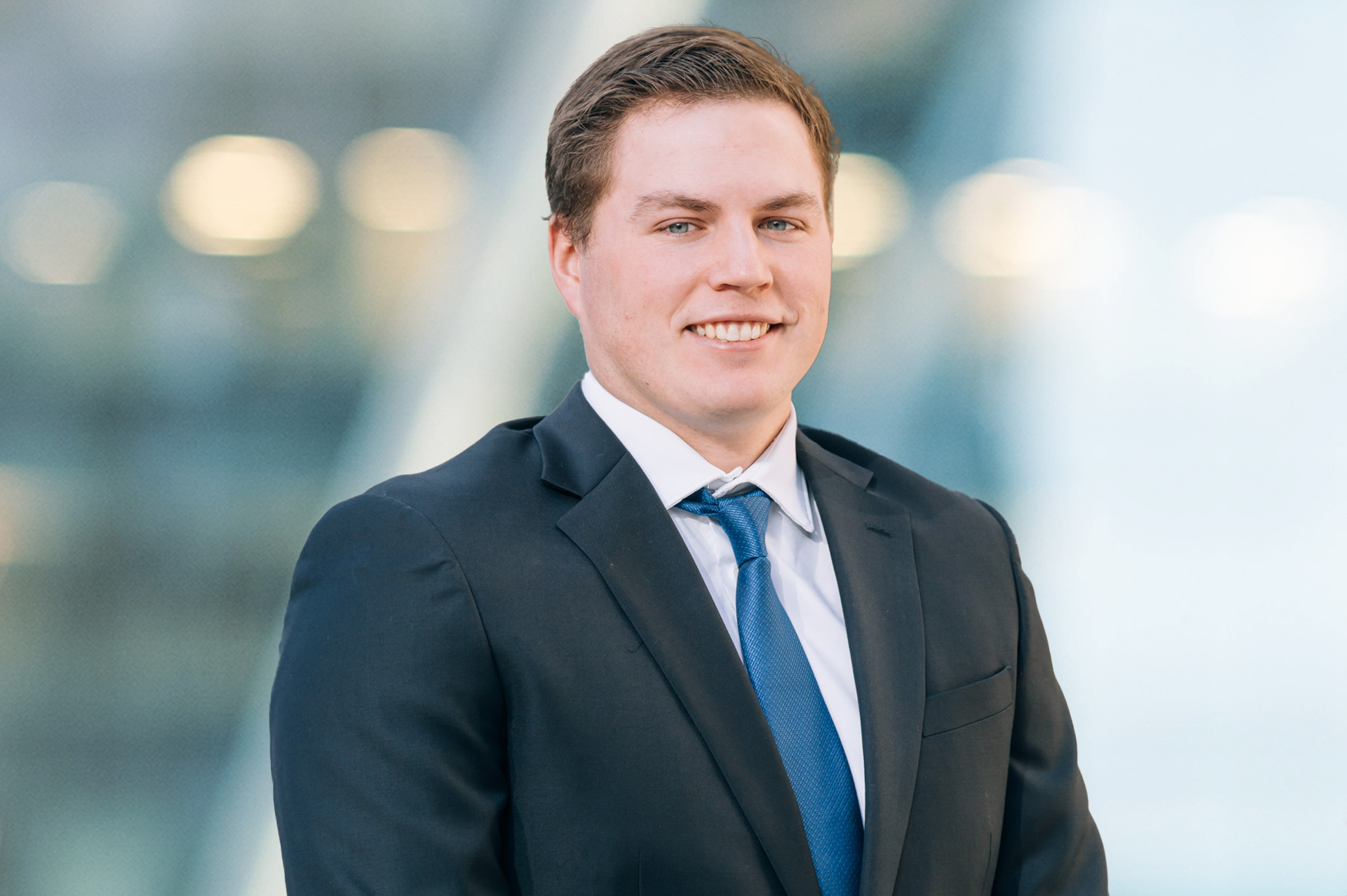 A young man in a dark suit, white shirt, and blue tie stands smiling in front of a blurred modern background with soft lighting.