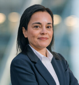 A woman with straight dark hair wearing a dark blazer and white shirt stands with arms crossed, smiling slightly. The background is blurred with soft lights, giving a professional, modern feel.