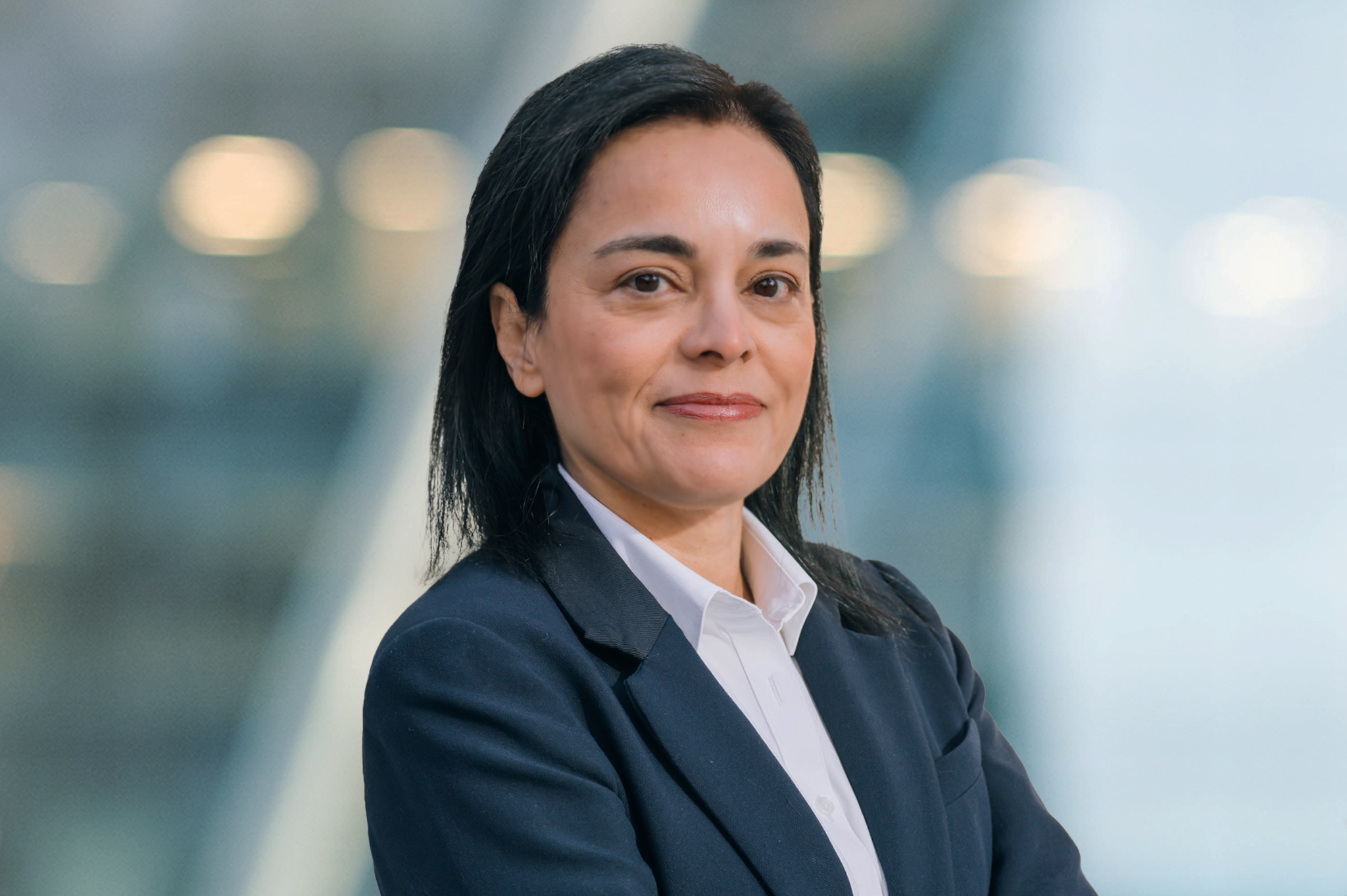 A woman with straight dark hair wearing a dark blazer and white shirt stands with arms crossed, smiling slightly. The background is blurred with soft lights, giving a professional, modern feel.