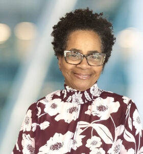A woman with short curly hair and glasses smiles at the camera. She is wearing a maroon and white floral blouse. The background is blurred with soft blue and light tones.