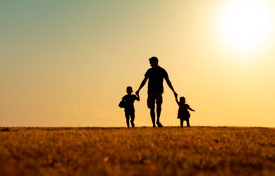 A silhouette of an adult holding hands with two young children, walking on grass at sunset, with a warm, orange sky in the background.