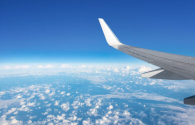 View from an airplane window showing a wingtip against a clear blue sky, with scattered white clouds over a vast landscape below.