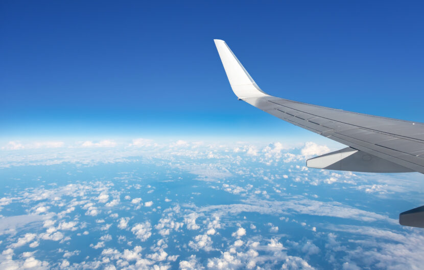 View from an airplane window showing a wingtip against a clear blue sky, with scattered white clouds over a vast landscape below.