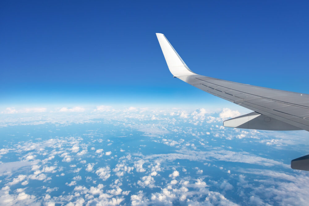 View from an airplane window showing a wingtip against a clear blue sky, with scattered white clouds over a vast landscape below.