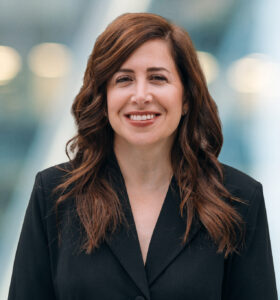 A woman with long, wavy brown hair wearing a black blazer smiles while standing in front of a blurred, modern indoor background with soft lighting.