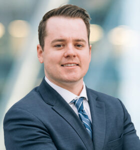 A man wearing a dark suit, white shirt, and striped tie stands with arms crossed, smiling confidently. The background is blurred with soft blue and white tones, suggesting a modern office setting.