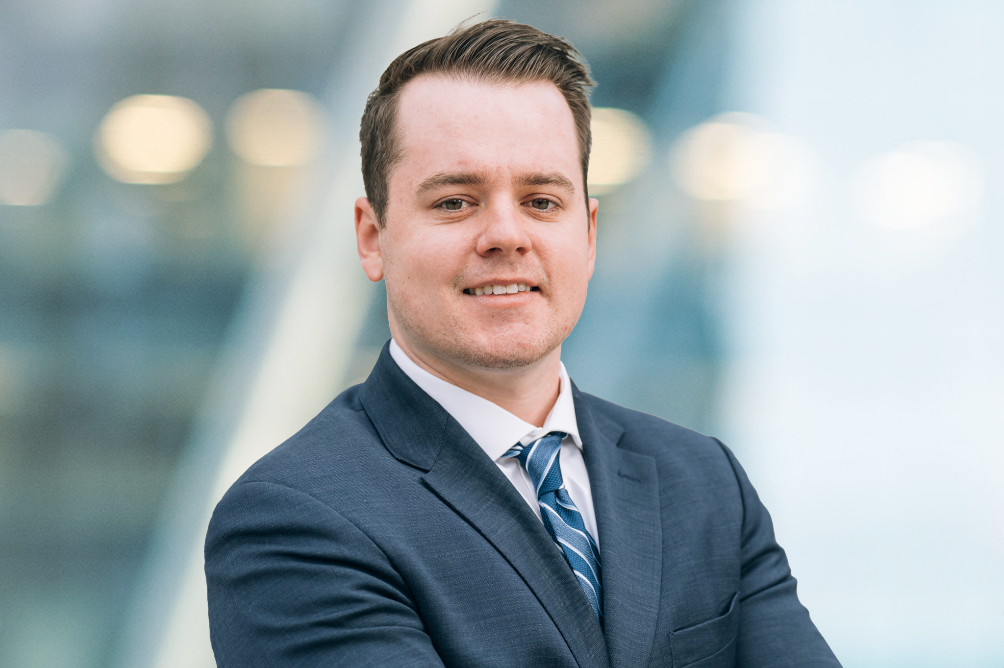 A man wearing a dark suit, white shirt, and striped tie stands with arms crossed, smiling confidently. The background is blurred with soft blue and white tones, suggesting a modern office setting.