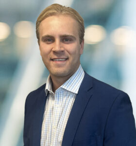 A smiling man with blond hair wearing a navy blue blazer and a light checkered shirt stands in front of a blurred background with soft lights.