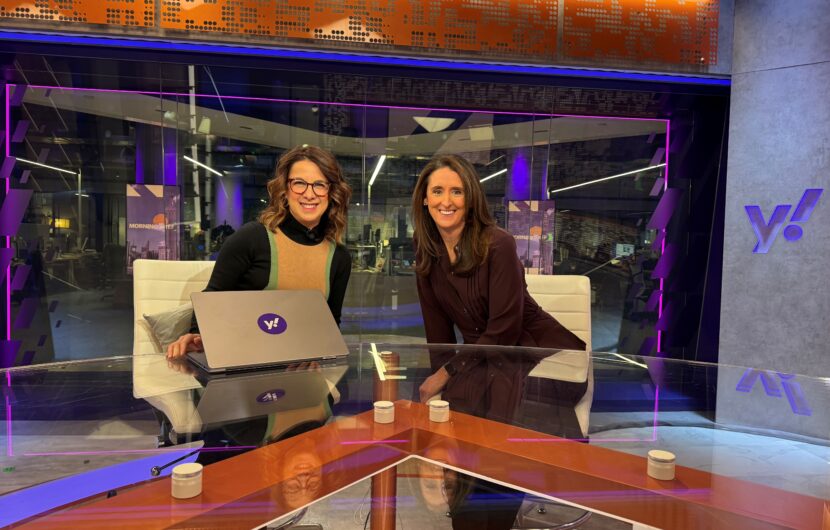 Two women sit at a modern news desk with a laptop displaying the Yahoo! logo. They are smiling at the camera, with purple and orange studio lighting and a Yahoo! logo visible in the background.
