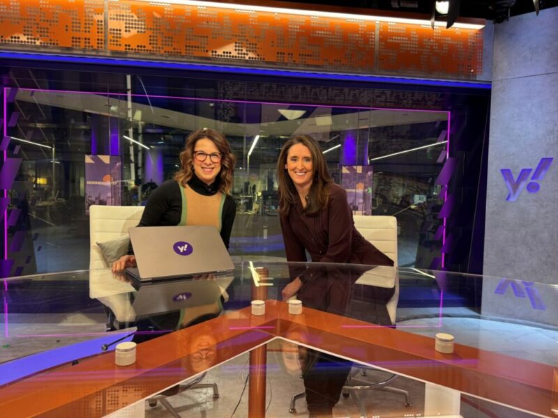 Two women sit at a modern news desk with a laptop displaying the Yahoo! logo. They are smiling at the camera, with purple and orange studio lighting and a Yahoo! logo visible in the background.