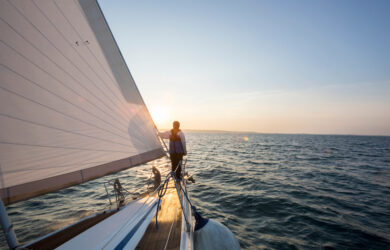 A person stands at the edge of a sailboat, holding onto the sail and looking out over the ocean as the sun sets, casting a warm glow on the water.