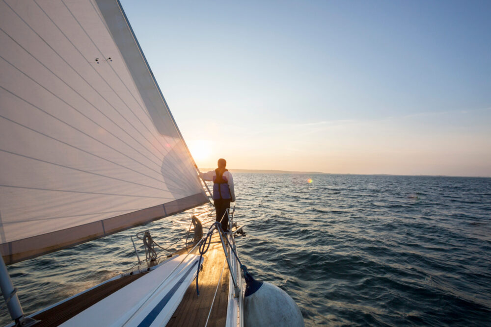A person stands at the edge of a sailboat, holding onto the sail and looking out over the ocean as the sun sets, casting a warm glow on the water.