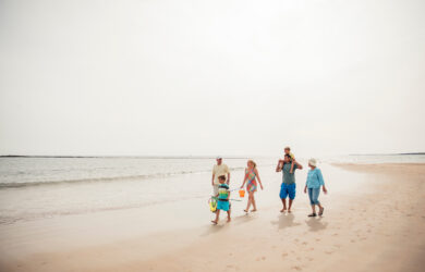 A family of five, including two children and three adults, walks along a sandy beach near the water on a bright, sunny day. The adults hold beach gear and the group appears happy and relaxed.