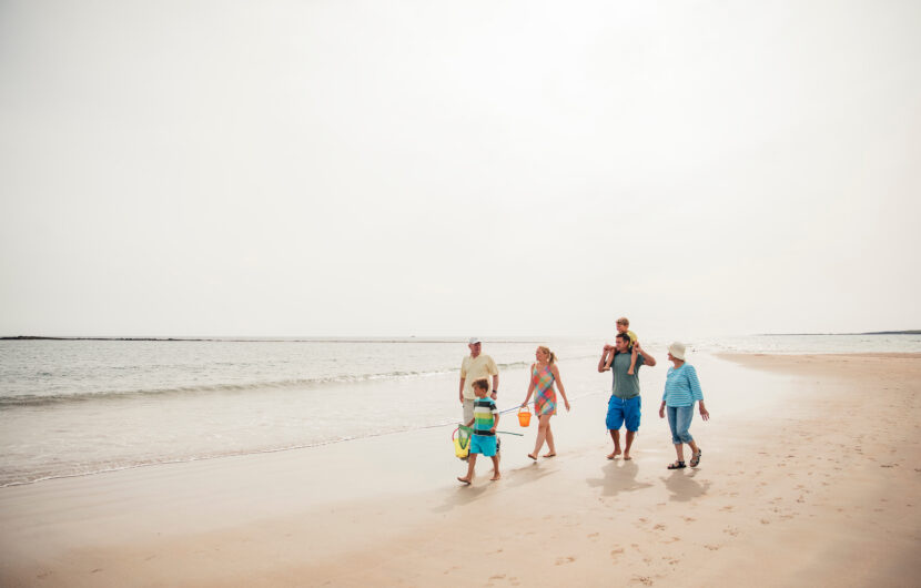 A family of five, including two children and three adults, walks along a sandy beach near the water on a bright, sunny day. The adults hold beach gear and the group appears happy and relaxed.