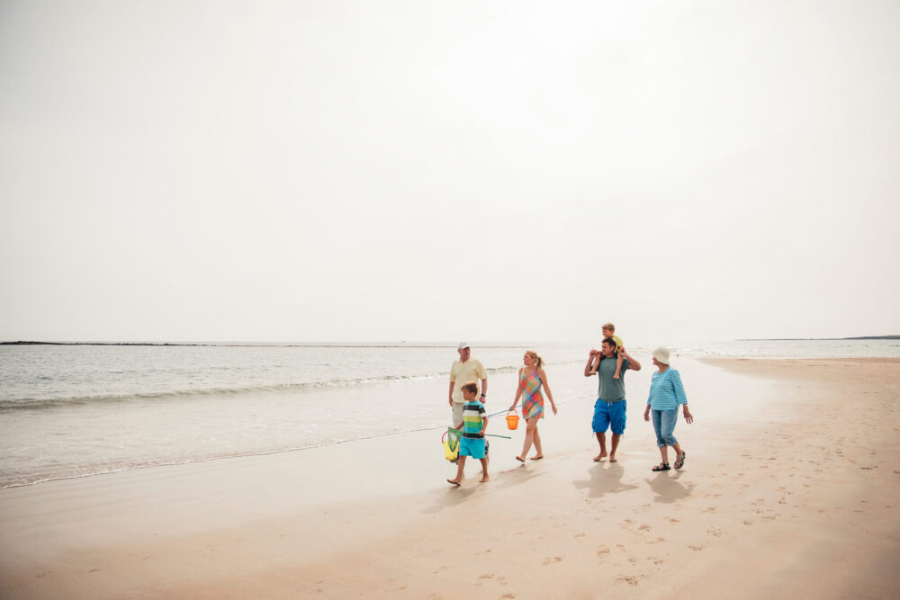 A family of five, including two children and three adults, walks along a sandy beach near the water on a bright, sunny day. The adults hold beach gear and the group appears happy and relaxed.