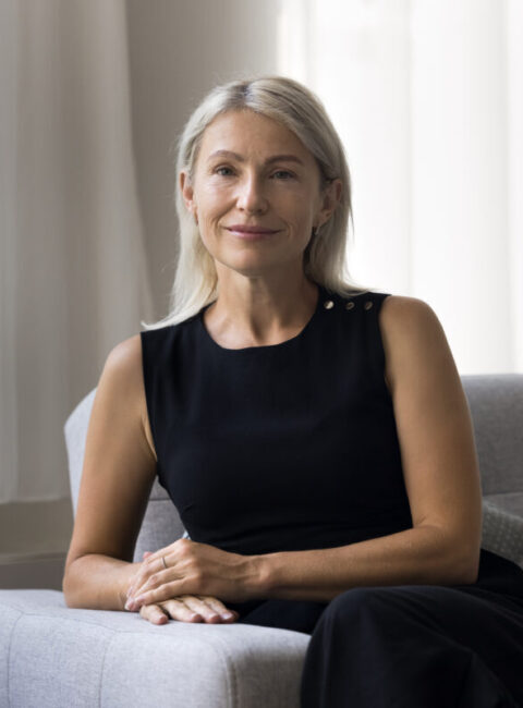 A woman with long blonde hair, wearing a sleeveless black dress, sits on a light grey armchair in a bright room with large windows and white curtains, smiling softly at the camera.