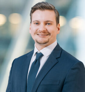 A man in a navy suit, white shirt, and dark tie is smiling at the camera. The background is blurred with soft lights and cool tones, giving a professional, modern atmosphere.