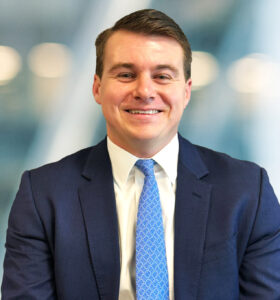 A man in a blue suit, white shirt, and blue patterned tie smiles while seated in front of a blurred office background.
