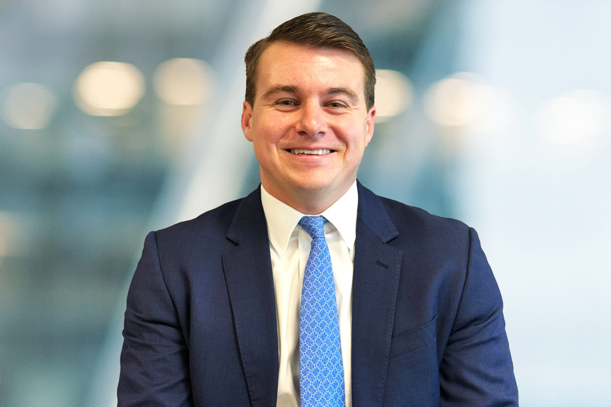 A man in a blue suit, white shirt, and blue patterned tie smiles while seated in front of a blurred office background.