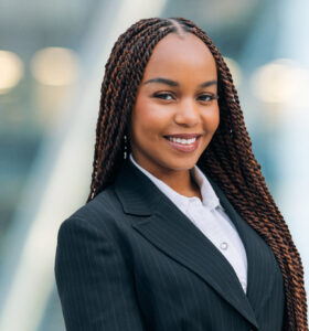 A young woman wearing a black pinstripe suit and white shirt smiles confidently at the camera. She has long, braided hair and is standing in front of a blurred, modern, glass-background.