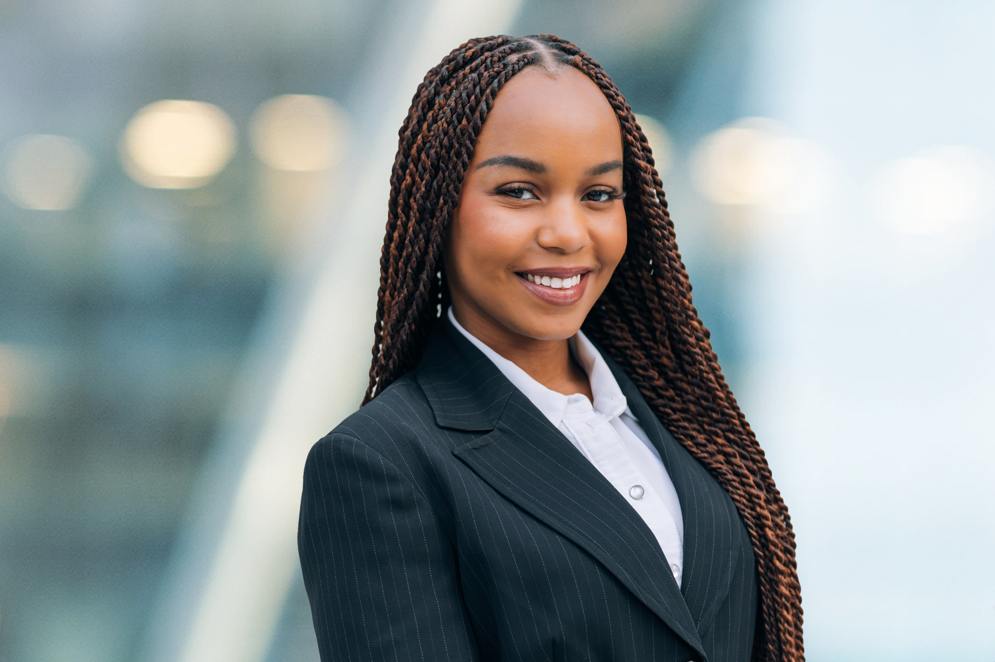 A young woman wearing a black pinstripe suit and white shirt smiles confidently at the camera. She has long, braided hair and is standing in front of a blurred, modern, glass-background.