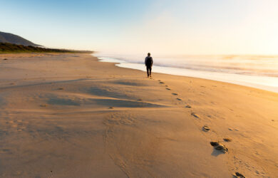 A person walks alone along a sandy beach at sunrise or sunset, leaving footprints behind. The sky is clear, and gentle waves roll onto the shore. A green hill lines the left edge of the beach in the distance.