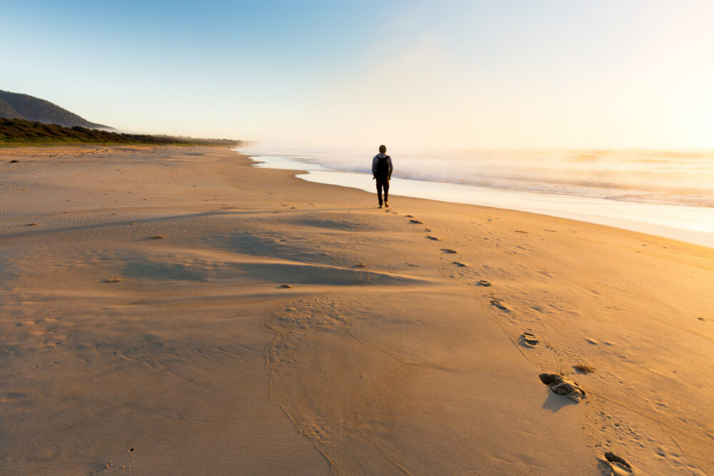 A person walks alone along a sandy beach at sunrise or sunset, leaving footprints behind. The sky is clear, and gentle waves roll onto the shore. A green hill lines the left edge of the beach in the distance.