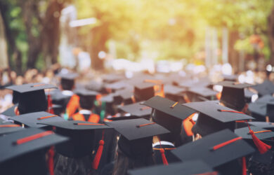 A large group of graduates wearing caps and gowns gather outdoors, facing forward. Sunlight filters through trees in the background, creating a celebratory and bright atmosphere.