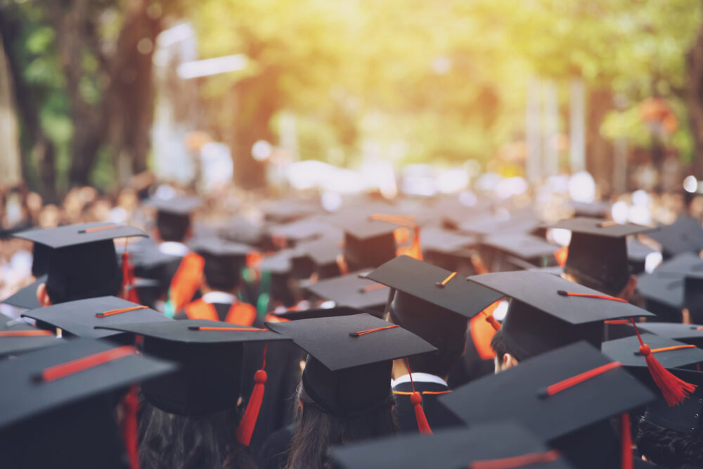 A large group of graduates wearing caps and gowns gather outdoors, facing forward. Sunlight filters through trees in the background, creating a celebratory and bright atmosphere.