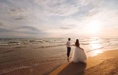 A bride and groom walk barefoot along a sandy beach at sunset, holding hands and facing the ocean. The sky is partly cloudy, and the sunlight creates a warm, romantic atmosphere.