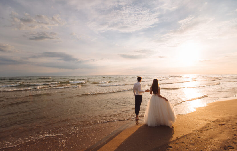 A bride and groom walk barefoot along a sandy beach at sunset, holding hands and facing the ocean. The sky is partly cloudy, and the sunlight creates a warm, romantic atmosphere.