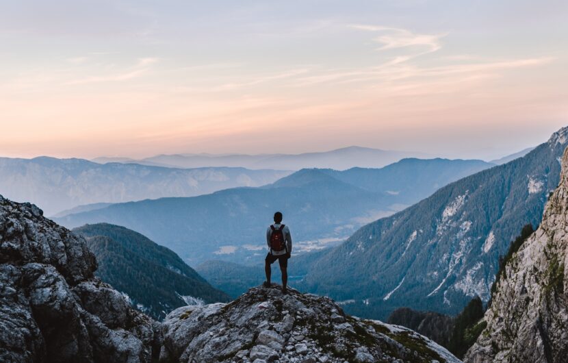 A person with a backpack stands on a rocky peak, overlooking a scenic valley and distant mountains at sunrise or sunset, with mist and layers of hills fading into the horizon under a colorful sky.